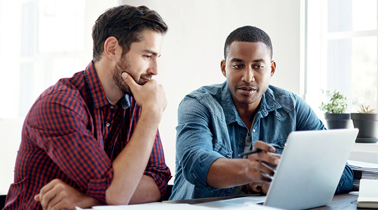 Two men are seated next to each other and are looking at the laptop in front of them